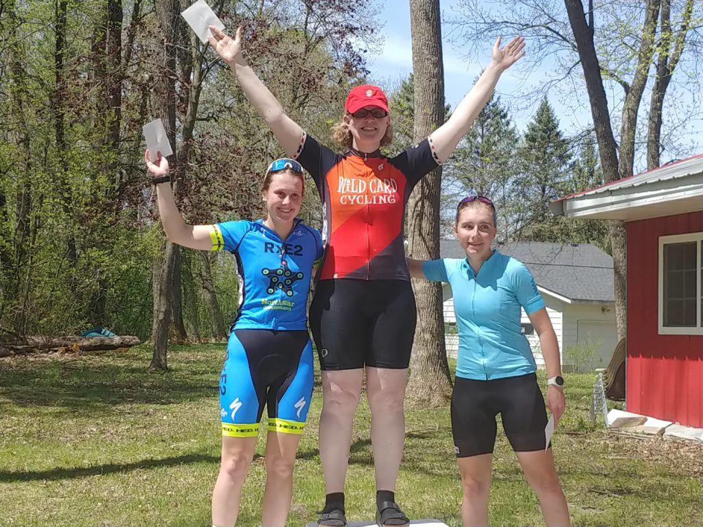 Three women on a ceremonial podium with jerseys as follows: Top step: Wild Card Cycling, Second step: North Star Development. Third step: light blue jersey without team designation